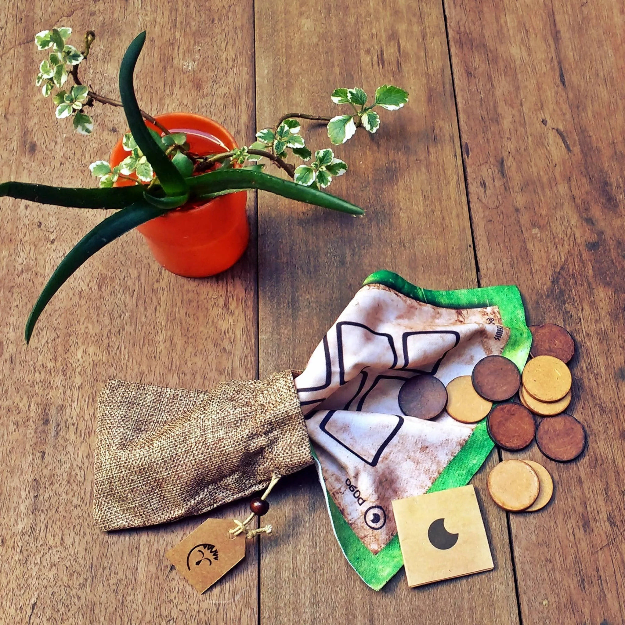 Tablero de tela del Pogo desplegado con sus fichas dispersas naturalmente, sale de su envoltorio que una bolsa de tela simil arpillera, vista cenital, una planta decora la mesa de madera